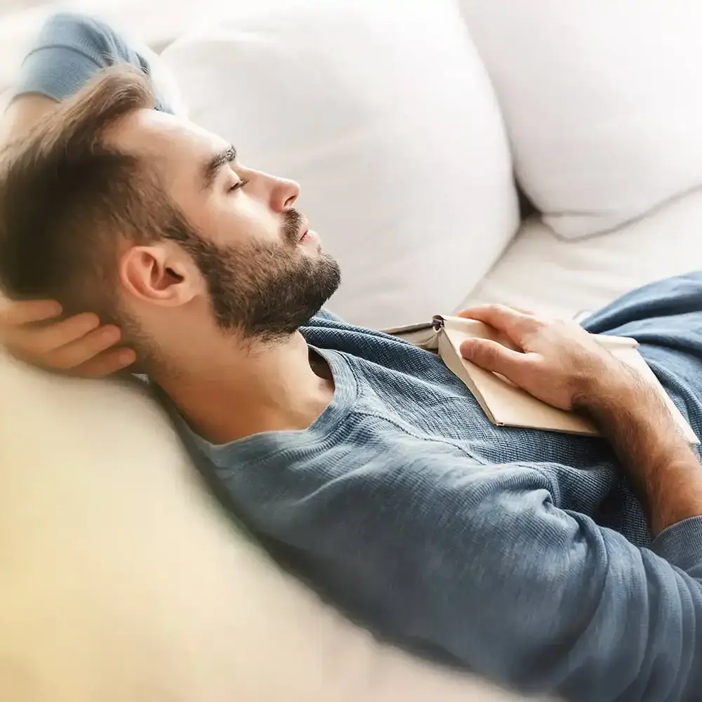 Mann mit geschlossenen Augen & Buch auf der Brust, der auf einem cremefarbenen Sofa entspannt.